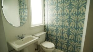 A small bathroom with a white sink, toilet, round mirror, and a window. One wall features blue and yellow leaf-patterned wallpaper, while the other walls are plain white.