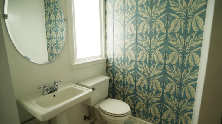A small bathroom with a white sink, toilet, round mirror, and a window. One wall features blue and yellow leaf-patterned wallpaper, while the other walls are plain white.