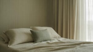 A neatly made bed with cream-colored sheets and pillows, featuring a small green cushion. Soft, beige curtains allow natural light to fill the calm, minimalist bedroom.