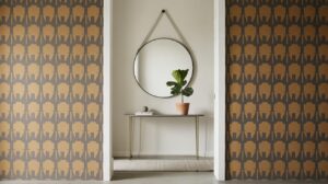 A modern entryway with bold geometric wallpaper, a slim console table holding a potted plant and books, and a large round mirror with a strap hanging above the table on a light-colored wall.